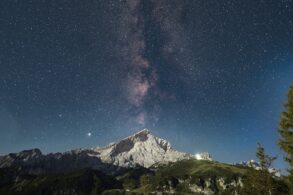 Über einem spitzen Berg inmitten einer Bergkette ist der Nachthimmel deutlich zu erkennen, die Milchstraße genau mittig über dem Gipfel.