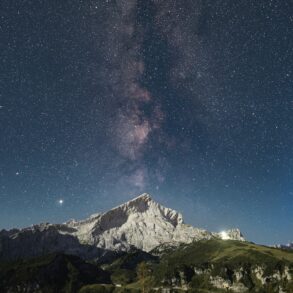 Über einem spitzen Berg inmitten einer Bergkette ist der Nachthimmel deutlich zu erkennen, die Milchstraße genau mittig über dem Gipfel.