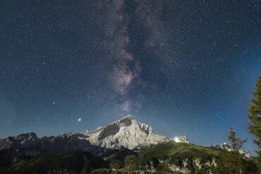 Über einem spitzen Berg inmitten einer Bergkette ist der Nachthimmel deutlich zu erkennen, die Milchstraße genau mittig über dem Gipfel.