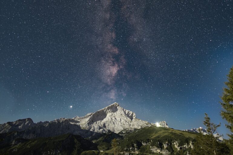 Über einem spitzen Berg inmitten einer Bergkette ist der Nachthimmel deutlich zu erkennen, die Milchstraße genau mittig über dem Gipfel.