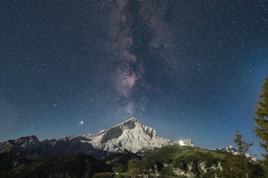 Über einem spitzen Berg inmitten einer Bergkette ist der Nachthimmel deutlich zu erkennen, die Milchstraße genau mittig über dem Gipfel.
