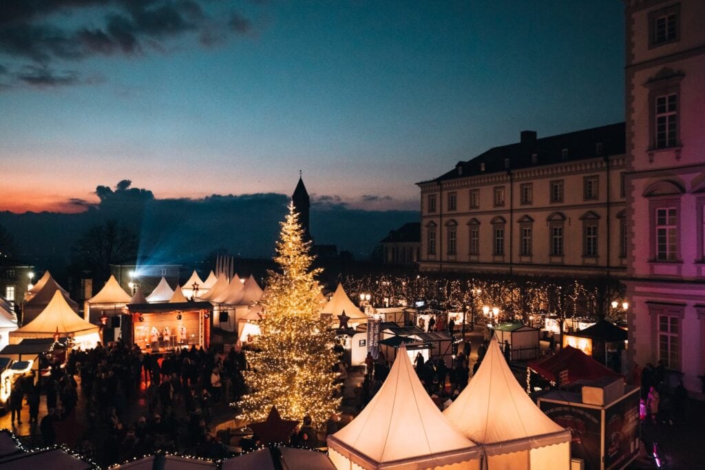 Ein Weihnachtsmarkt mit von innen erleuchteten Zelten spät am Abend. Im Zentrum des Marktes steht ein hell leuchtender Weihnachtsbaum. Auf der rechten Bildseite ragt ein dreigeschossiges Schloss ins Bild, vor dem der Weihnachtsmarkt aufgebaut ist.
