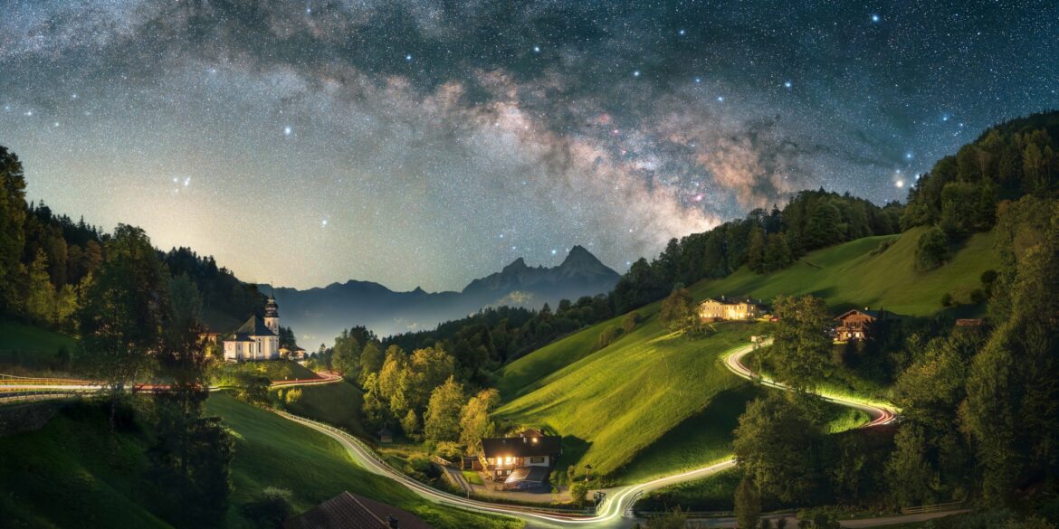 A long exposure of a Bavarian valley at night. On a hill to the left stands a small chapel, a road from left to right is illuminated by long drawn car lights, to the right are some houses. The peaks of the Alps are rising up in the background, the night sky with a clearly visible Milky Way crowns the image.