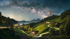 A long exposure of a Bavarian valley at night. On a hill to the left stands a small chapel, a road from left to right is illuminated by long drawn car lights, to the right are some houses. The peaks of the Alps are rising up in the background, the night sky with a clearly visible Milky Way crowns the image.