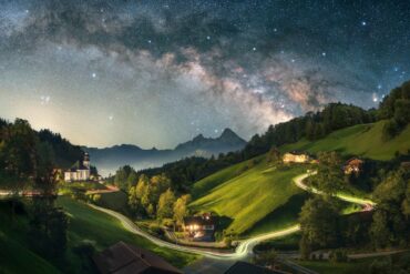 A long exposure of a Bavarian valley at night. On a hill to the left stands a small chapel, a road from left to right is illuminated by long drawn car lights, to the right are some houses. The peaks of the Alps are rising up in the background, the night sky with a clearly visible Milky Way crowns the image.