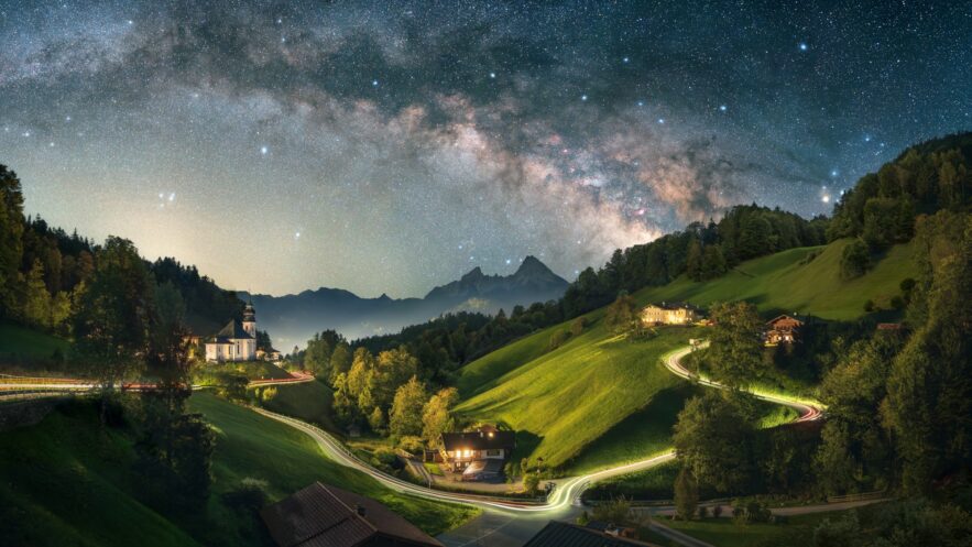 A long exposure of a Bavarian valley at night. On a hill to the left stands a small chapel, a road from left to right is illuminated by long drawn car lights, to the right are some houses. The peaks of the Alps are rising up in the background, the night sky with a clearly visible Milky Way crowns the image.