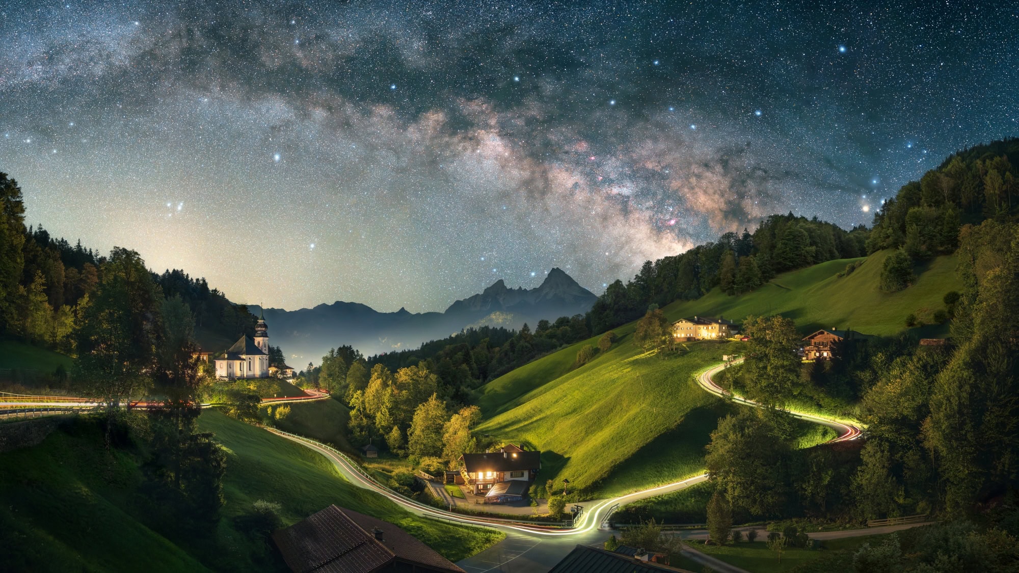A long exposure of a Bavarian valley at night. On a hill to the left stands a small chapel, a road from left to right is illuminated by long drawn car lights, to the right are some houses. The peaks of the Alps are rising up in the background, the night sky with a clearly visible Milky Way crowns the image.