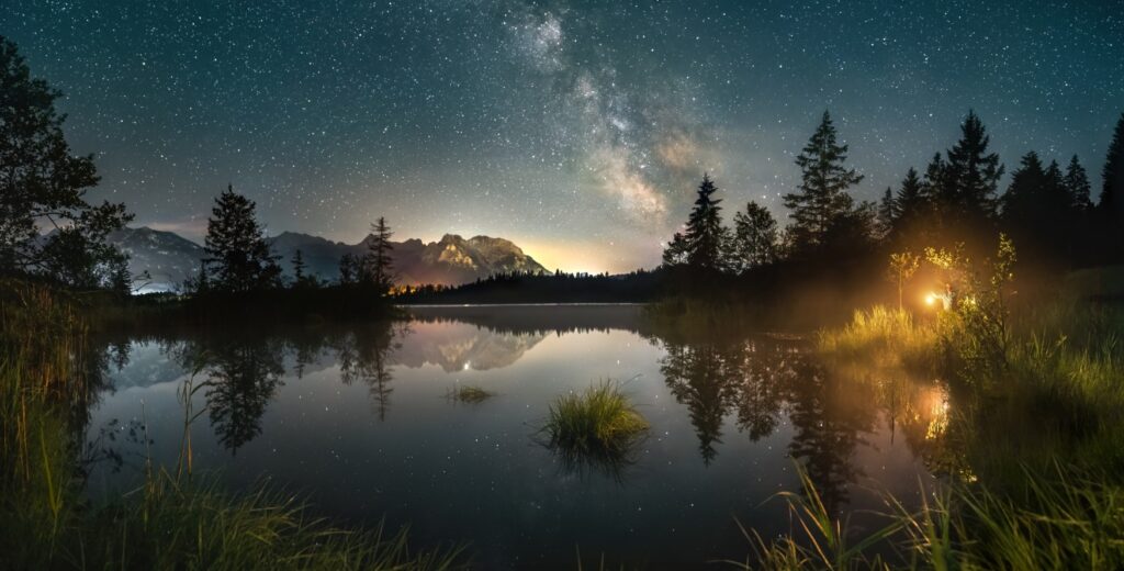 Ein still liegender Alpensee bei Nacht; über dem See sieht man klar die Milchstraße. Links im Hintergrund zeichnen sich die Alpen ab, rechts am Seeufer steht ein Mann mit einer gelb leuchtenden Laterne, deren Schein sich auf der Wasseroberfläche spiegelt.