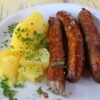 Auf einem weißen Teller liegen links einige Salzkartoffeln mit Petersilie und rechts drei rötliche, grobe Bratwürste. Unter dem Teller ist eine Speisekarte erkennbar.