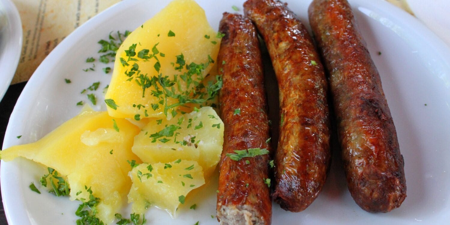 Auf einem weißen Teller liegen links einige Salzkartoffeln mit Petersilie und rechts drei rötliche, grobe Bratwürste. Unter dem Teller ist eine Speisekarte erkennbar.