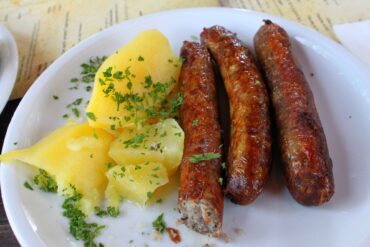 Auf einem weißen Teller liegen links einige Salzkartoffeln mit Petersilie und rechts drei rötliche, grobe Bratwürste. Unter dem Teller ist eine Speisekarte erkennbar.