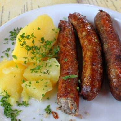 Auf einem weißen Teller liegen links einige Salzkartoffeln mit Petersilie und rechts drei rötliche, grobe Bratwürste. Unter dem Teller ist eine Speisekarte erkennbar.