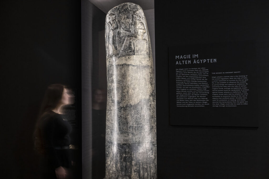 Eine etwa zwei Meter hohe, schwarze Säule mit eingravierter Keilschrift, angeleuchtet in einer Museumsvitrine. Auf dem obersten Abschnitt der Säule sind ein stehender und ein sitzender Mann zu erkennen. An der schwarzen Wand rechts hinter der Säule lautet eine Überschrift "Magie im alten Ägypten", darunter ein Infotext auf englisch und auf deutsch. Links von der Säule steht eine Frau in schwarzer Kleidung und schaut auf das Exponat.