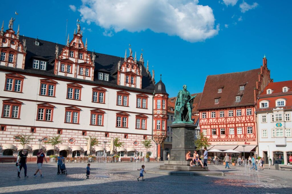 On a medieval square with colorful, magnificent half-timbered houses stands a copper statue of a nobleman. He holds a scroll in his left hand.