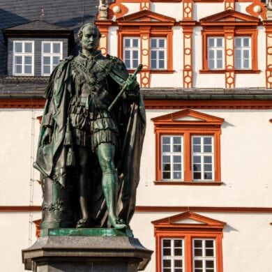 On a medieval square with colorful, magnificent half-timbered houses stands a copper statue of a nobleman. He holds a scroll in his left hand.