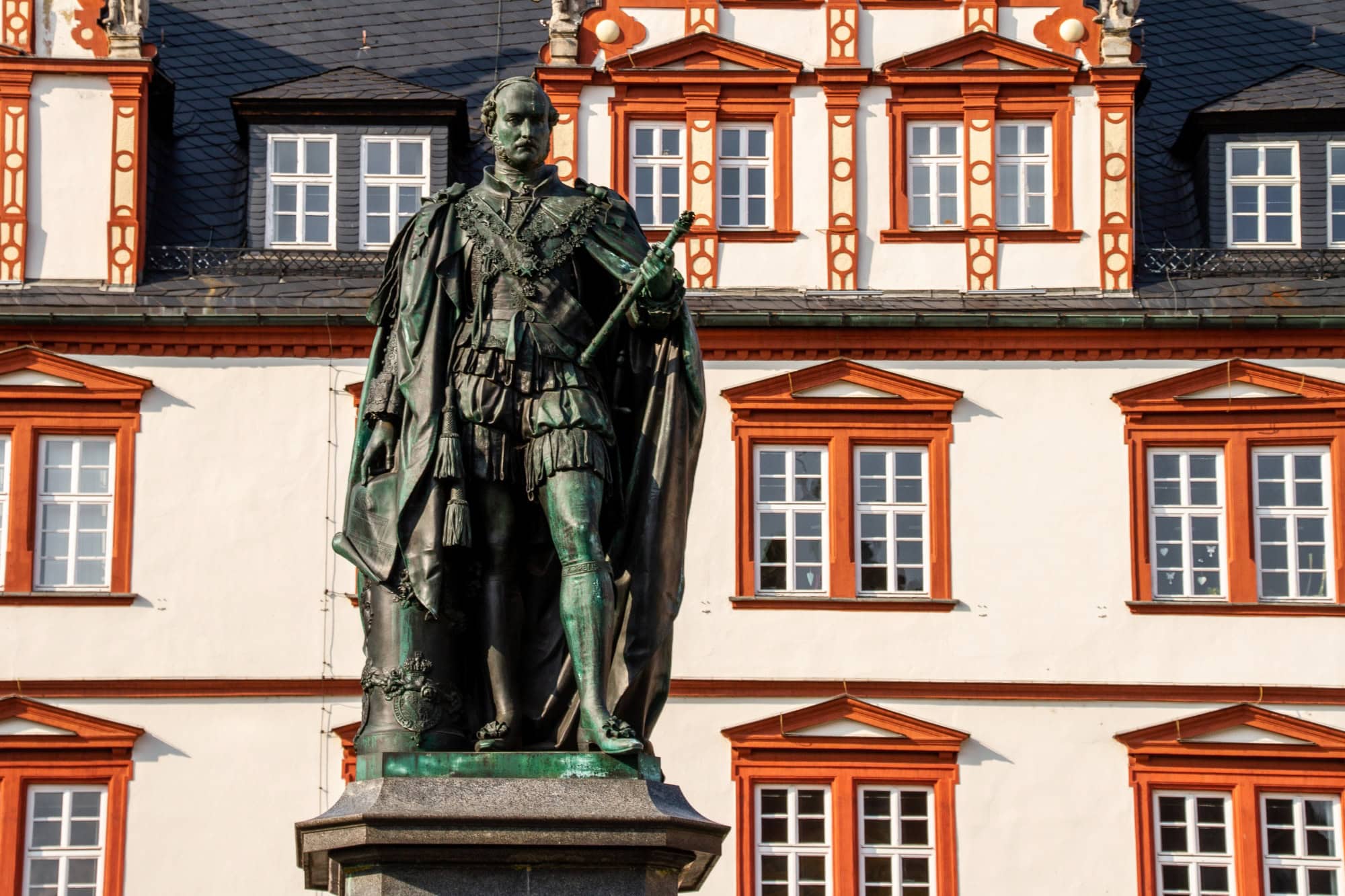 On a medieval square with colorful, magnificent half-timbered houses stands a copper statue of a nobleman. He holds a scroll in his left hand.