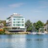 An einem Flussufer steht ein moderner, eckiger Bau mit großen Fenstern, drei Stockwerke hoch, obenauf ein Penthouse. Zum Ufer zeigt eine steinerne Terrasse. Rechts grenzt am Ufer ein kleiner Yachthafen an, dahinter viele Bäume und einige Hausdächer. Über allem strahlend blauer Himmel.