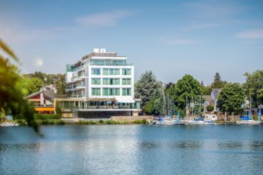 An einem Flussufer steht ein moderner, eckiger Bau mit großen Fenstern, drei Stockwerke hoch, obenauf ein Penthouse. Zum Ufer zeigt eine steinerne Terrasse. Rechts grenzt am Ufer ein kleiner Yachthafen an, dahinter viele Bäume und einige Hausdächer. Über allem strahlend blauer Himmel.