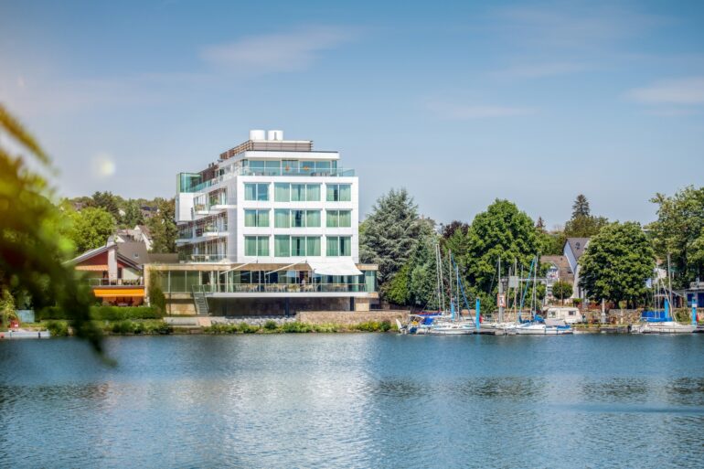 An einem Flussufer steht ein moderner, eckiger Bau mit großen Fenstern, drei Stockwerke hoch, obenauf ein Penthouse. Zum Ufer zeigt eine steinerne Terrasse. Rechts grenzt am Ufer ein kleiner Yachthafen an, dahinter viele Bäume und einige Hausdächer. Über allem strahlend blauer Himmel.