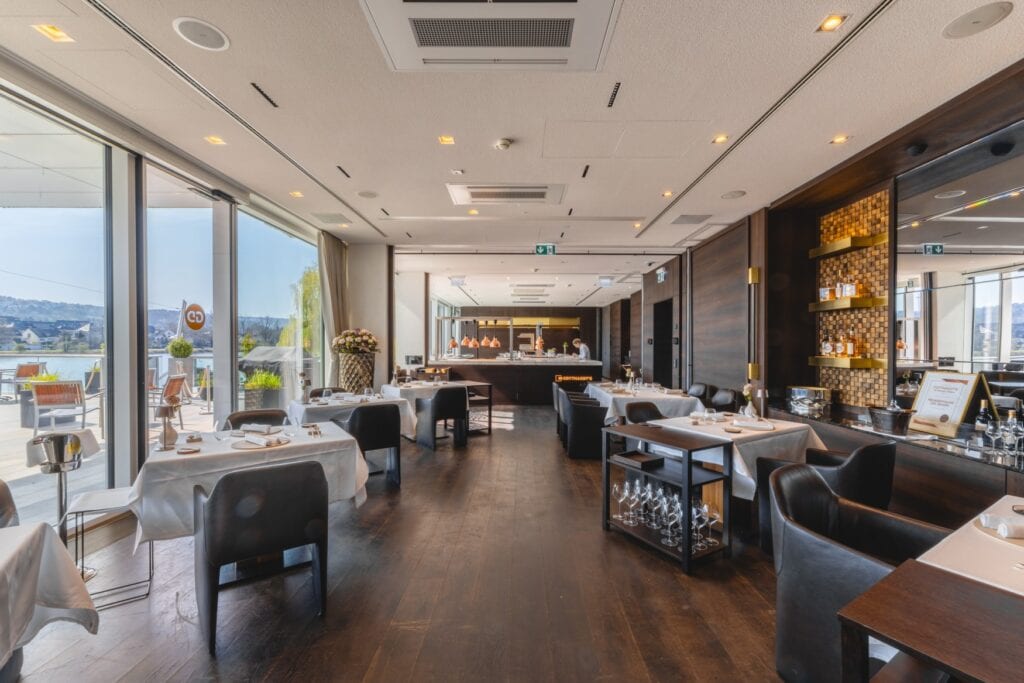 Speisesaal in einem gehobenen Restaurant; vor einer Fensterfront links stehen drei quadratische Tische mit weißen Decken und jeweils zwei Stühlen, daneben je ein Weinkühler. Auf der rechten Seite nochmals zwei einzelne Tische, weiter hinten im Saal ein längerer Tisch für vier Personen. Ganz im Hintergrund ist ein offener Bereich zum Anrichten zu sehen, rechts im Saal eine Bar mit großen Spiegel. Durch die Fensterfront links kann man hinter einer Terrasse einen Fluss erkennen.