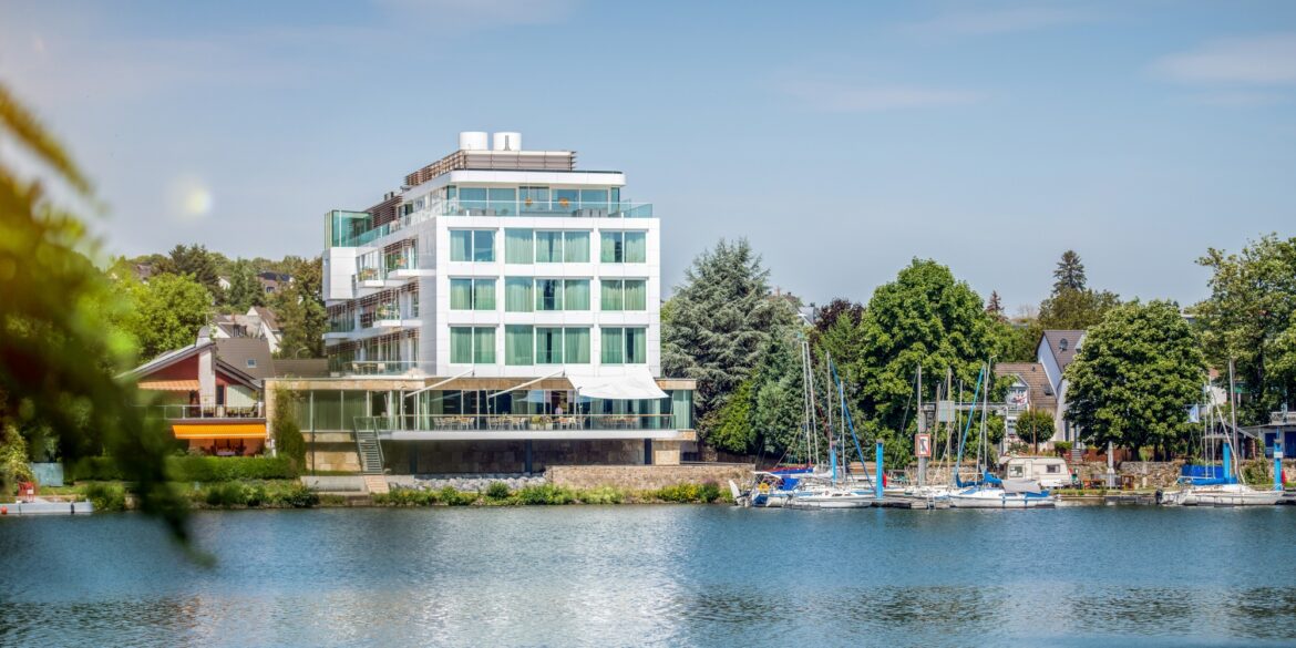 On the bank of a river stands a modern, angular building with large windows, three stories high, topped by a penthouse. A stone terrace faces the riverbank. To the right, a small marina borders the riverbank, behind it many trees and a few rooftops. Above it all, a bright blue sky.
