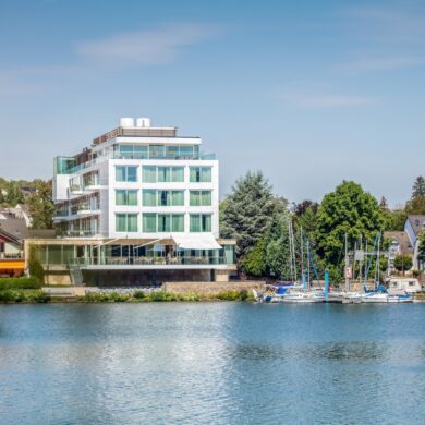 On the bank of a river stands a modern, angular building with large windows, three stories high, topped by a penthouse. A stone terrace faces the riverbank. To the right, a small marina borders the riverbank, behind it many trees and a few rooftops. Above it all, a bright blue sky.
