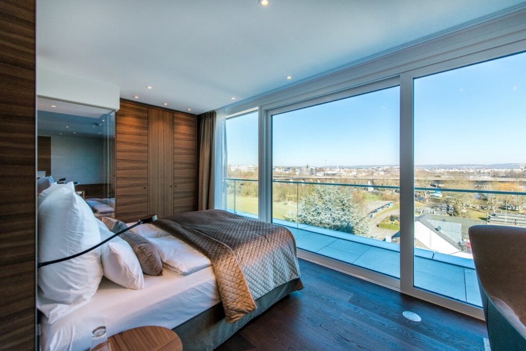 Ein modernes Hotelzimmer; links ein breites Bett mit vielen Kissen und braun-goldener Tagesdecke, dahinter ein mit Glaswand abgetrenntes Bad, rechts davon ein brauner Wandschrank. Das Bett ist auf die Fensterfront zur Rechten ausgerichtet, dahinter ist ein grüner Park mit herbstlichen Bäumen zu erkennen. Der Himmel strahlt blau.
