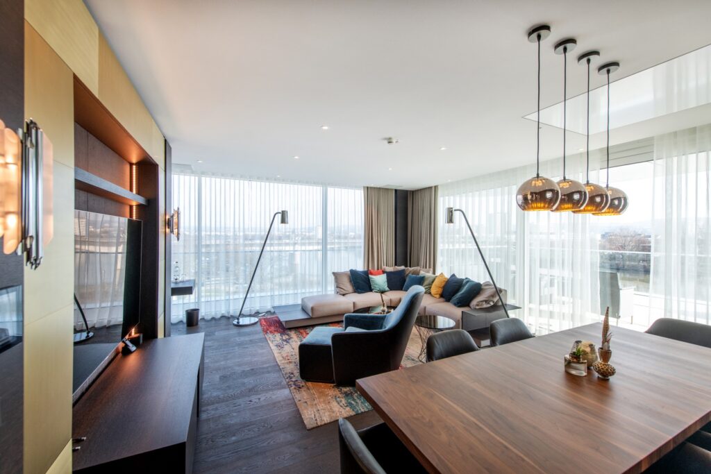 Living area in a hotel suite. On the left side of the picture is a wall with gold paneling, into which a large television is embedded. On the right side at the bottom is a wooden table with two leather armchairs, and behind it is an upholstered seating area with many colorful cushions and a freestanding upholstered armchair, both on a carpet. The floor consists of dark floorboards. The right and rear walls of the room are completely glazed, allowing daylight to enter.