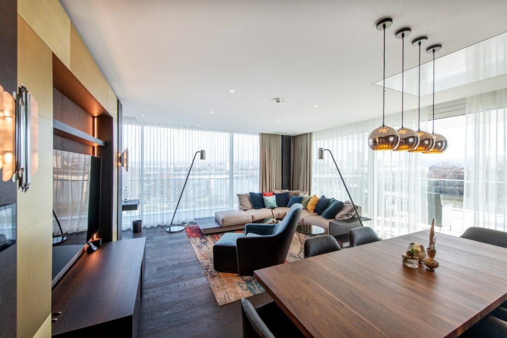 Wohnbereich in einer Hotelsuite. Links im Bild eine Wand mit goldener Verkleidung, in die ein großer Fernseher eingelassen ist. Rechts am unteren Rand ein Holztisch mit zwei Ledersesseln, rechts dahitner eine gepolsterte Sitzecke mit vielen bunten Kissen und einem freistehenden Polstersessel, beides auf einem Teppich. Der Boden besteht aus dunklen Dielen. Die rechte und hintere Wand des Raums sind komplett verglast, es fällt Tageslicht hinein.