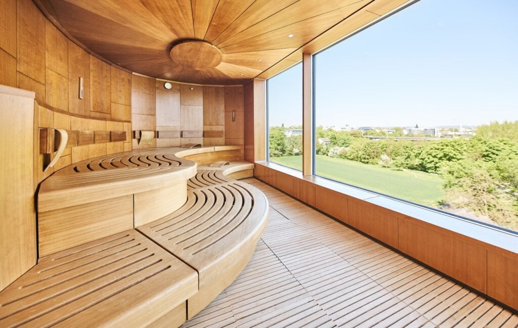 Eine moderne, hölzerne Sauna. Auf der linken Seite sind zwei Sitzebenen S-förmig geschwungen, auf der rechten Seite gewährt ein breites, bodentiefes Fenster den Blick auf einen grünen Park unter blauem Himmel.