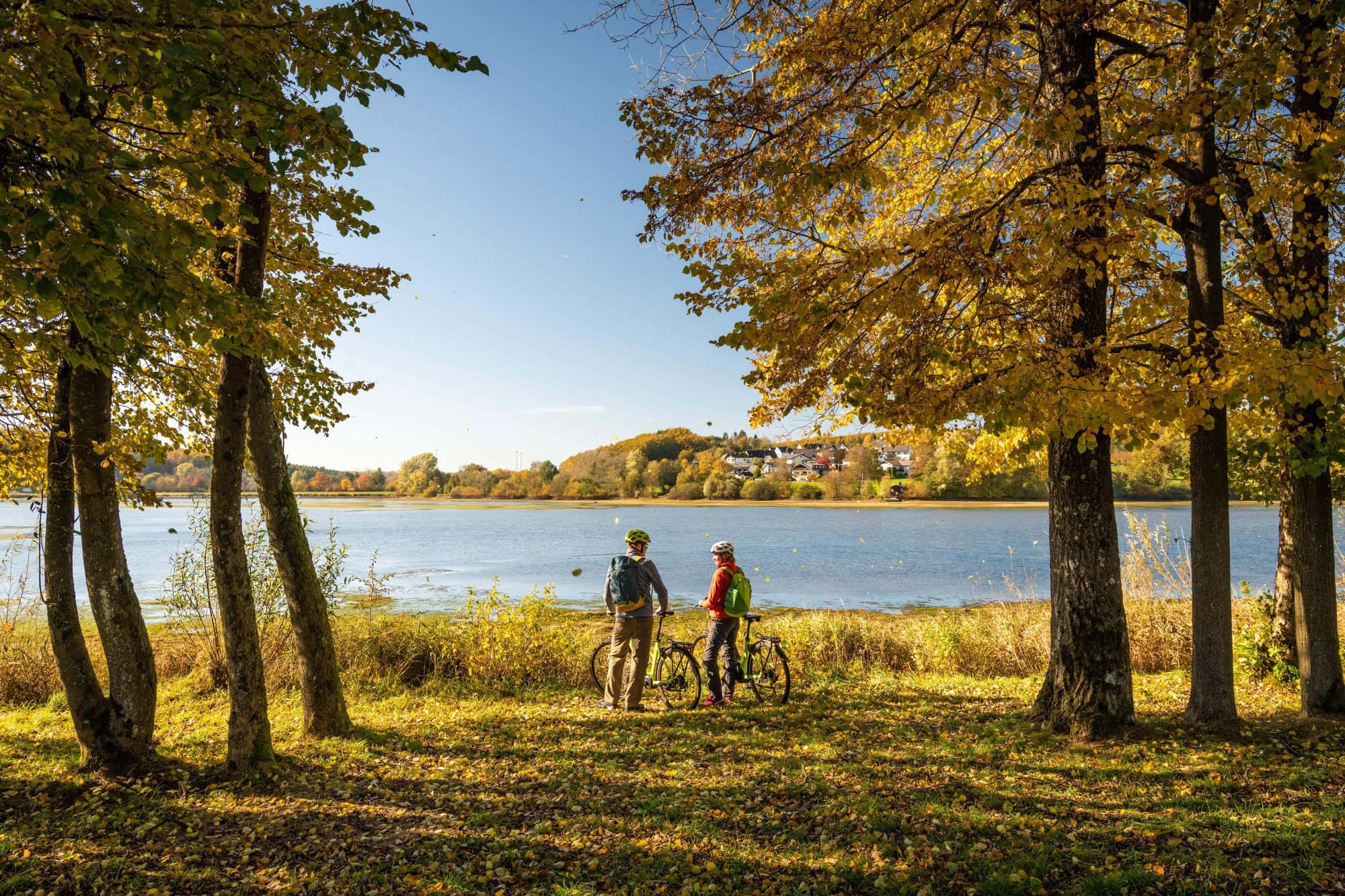 Two people in fall outdoor clothing stand with their backs to the viewer next to their bicycles on the green shore of a lake. To their left and right are several trees with colorful fall leaves. The sun shines on the lake, and a small village can be seen between the trees on the shore behind them.