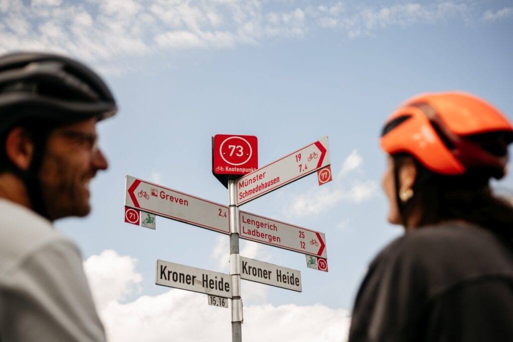 Ein Wegweiser, obenauf ein rotes Schild mit der Aufschrift "73 Knotenpunkt"; darunter gehen mehrere Richtungsweiser ab, die unter anderem nach Münster, Greven und Lengerich zeigen. Zuunterst ein Straßenschild, das den Weg als "Kroner Heide" auszeichnet. Links und rechts unscharf im Vordergrund ein Mann und eine Frau mit Fahrradhelmen.