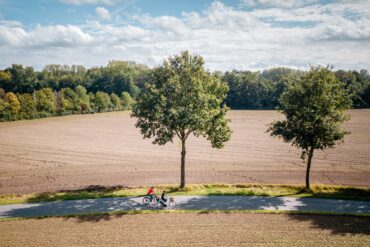 Drohnenaufnahme von zwei Radfahrer*innen auf einem Landweg mitten durch zwei Felder, von linsk nach rechts. Die Felder wirken frisch abgeerntet, am Wegesrand stehen zwei junge Bäume. Hinter dem hinteren Feld grenzt ein dichter Wald an, darüber blauer Himmel mit einigen Wolken.