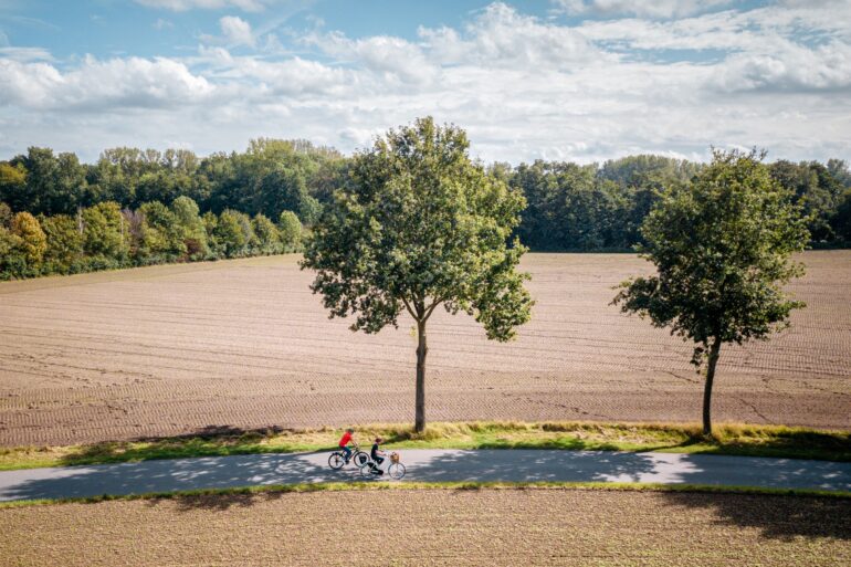 Drohnenaufnahme von zwei Radfahrer*innen auf einem Landweg mitten durch zwei Felder, von linsk nach rechts. Die Felder wirken frisch abgeerntet, am Wegesrand stehen zwei junge Bäume. Hinter dem hinteren Feld grenzt ein dichter Wald an, darüber blauer Himmel mit einigen Wolken.