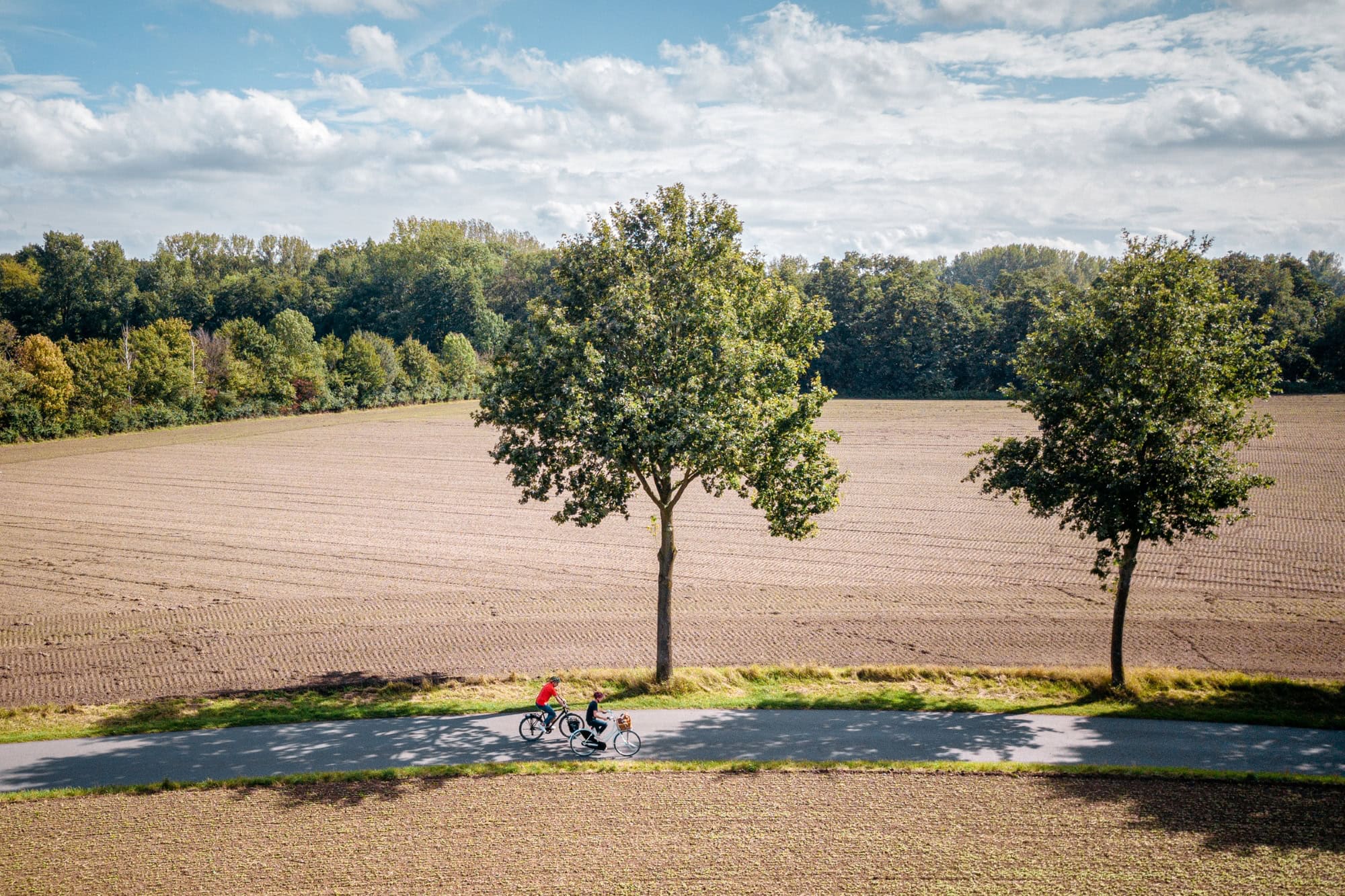 Drohnenaufnahme von zwei Radfahrer*innen auf einem Landweg mitten durch zwei Felder, von linsk nach rechts. Die Felder wirken frisch abgeerntet, am Wegesrand stehen zwei junge Bäume. Hinter dem hinteren Feld grenzt ein dichter Wald an, darüber blauer Himmel mit einigen Wolken.