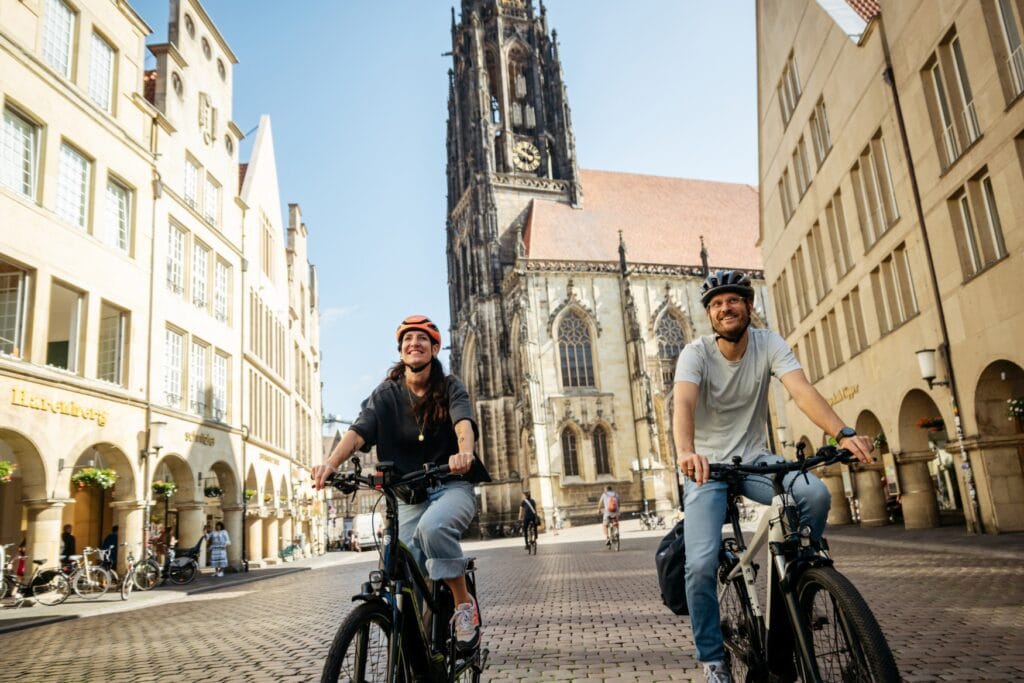 Ein Mann und eine Frau fahren auf Fahrrädern links und rechts am Betrachter vorbei auf einer Pflasterstraße. Links reihen sich die historischen Fassaden des Prinzipalmarkts in Münster aneinander, rechts im Hintergrund erhebt sich die gotische Lambertikirche in die Höhe. Über allem blauer Himmel.