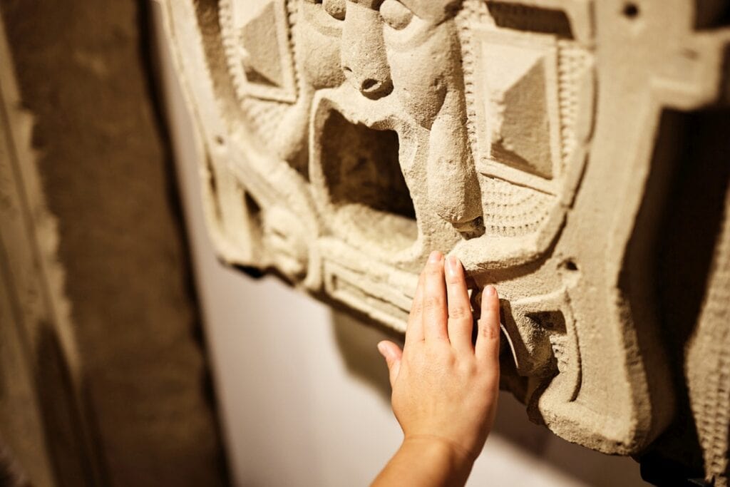 Detailaufnahme einer Hand, die ein Relief aus Sandstein, das an einer Wand hängt, berührt. Das Relief zeigt ein stilisiertes Menschengesicht mit weit aufgerissenem Mund, vielleicht als Wasserspeier gedacht.