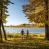 Zwei Personen in herbstlicher Outdoorkleidung stehen, die Rücken zum Betrachter, neben ihren Fahrrädern an einem grünen Seeufer. Links und rechts von ihnen stehen mehrere Bäume mit herbstbunten Blättern. Auf den See scheint die Sonne, am Ufer dahinter ist zwischen Bäumen eine kleine Ortschaft zu sehen.