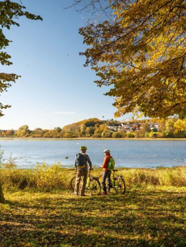 Zwei Personen in herbstlicher Outdoorkleidung stehen, die Rücken zum Betrachter, neben ihren Fahrrädern an einem grünen Seeufer. Links und rechts von ihnen stehen mehrere Bäume mit herbstbunten Blättern. Auf den See scheint die Sonne, am Ufer dahinter ist zwischen Bäumen eine kleine Ortschaft zu sehen.