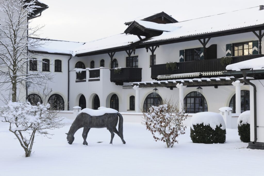 Innehof eines weiß getünchten Guthofs im Winter. Alles liegt unter einer dichten Schneedecke, im Zentrum des Hofs steht eine ebenfalls verschneite Bronze eines grasenden Pferds