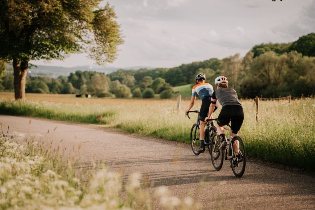 Two cyclists in sportswear are riding away from the viewer on a narrow asphalt path to the left. The path leads through a green field, with a single deciduous tree standing on the right-hand side of the road. A wooded hilly landscape can be seen blurred in the background on the right.