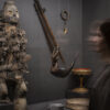 Vitrinie in einer Ausstellung. Im Fokus steht eine stilisierte, hölzerne Statue eines Menschen, in die zahllose Klingen, Beile und andere Metallstücke eingelassen sind. Rechts davon hängt ein verziertes Tierhorn von der Decke, an der Wand dahinter hängt ein Amulett mit einen spitzen Tierzahn.