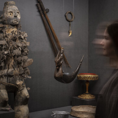 Vitrinie in einer Ausstellung. Im Fokus steht eine stilisierte, hölzerne Statue eines Menschen, in die zahllose Klingen, Beile und andere Metallstücke eingelassen sind. Rechts davon hängt ein verziertes Tierhorn von der Decke, an der Wand dahinter hängt ein Amulett mit einen spitzen Tierzahn.