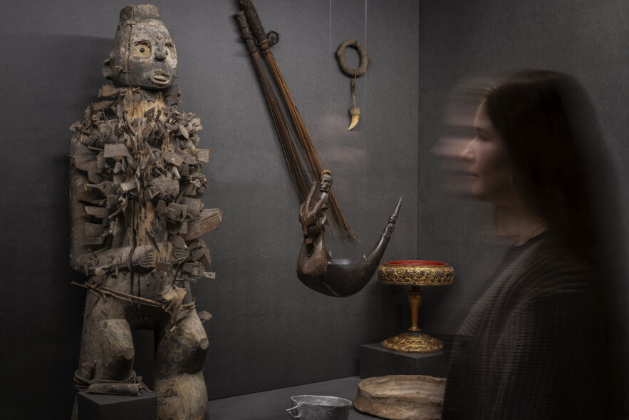 Vitrinie in einer Ausstellung. Im Fokus steht eine stilisierte, hölzerne Statue eines Menschen, in die zahllose Klingen, Beile und andere Metallstücke eingelassen sind. Rechts davon hängt ein verziertes Tierhorn von der Decke, an der Wand dahinter hängt ein Amulett mit einen spitzen Tierzahn.