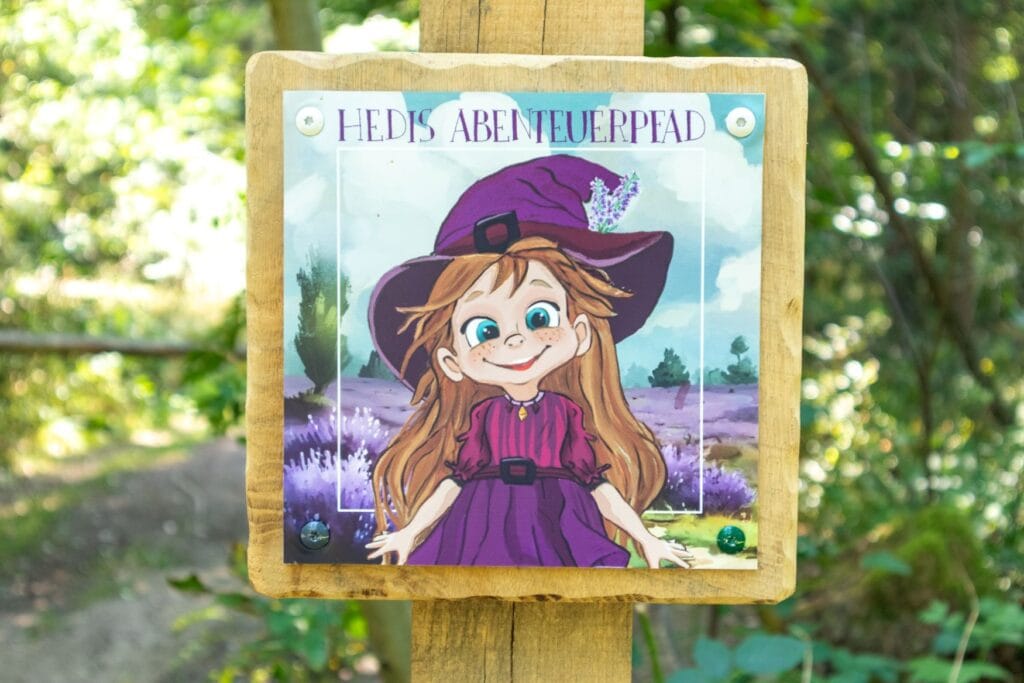 Ein quadratisches Schild in einem Wald zeigt in Nahaufnahme ein junges, lächelndes Mädchen in einem lila Kleid und mit einem lila Spitzhut. Darüber steht "HEDIS ABENTEUERPFAD".