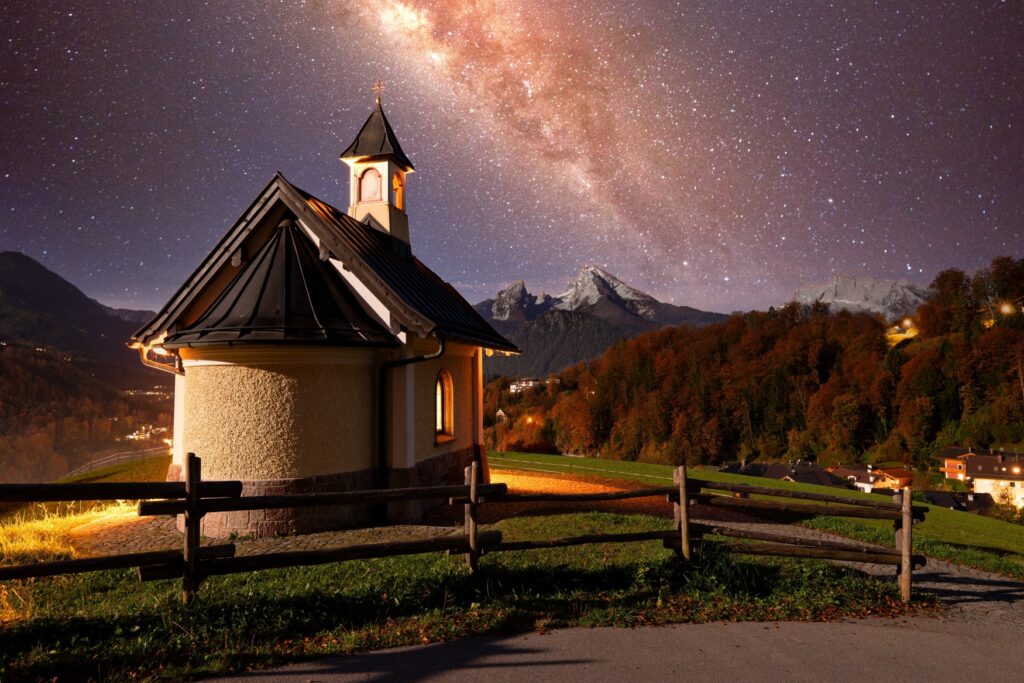 Panoramaaufnahme einer kleinen Kapelle in den bayerischen Alpen bei Nacht; die Kapelle steht auf einer Hügelkuppe, im Hintergrund öffnet sich ein Tal mit einigen Ortschaften, dahinter ragen die Gipfel der Alpen auf. Über allem der nächtliche Himmel mit deutlich sichtbarer Milchstraße.