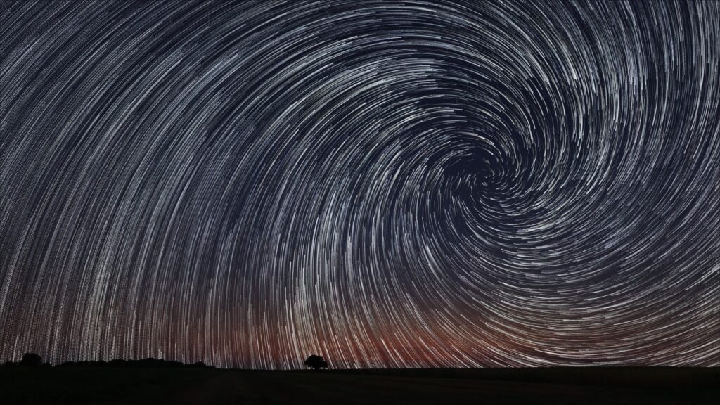 Wide-angle long exposure shot of the night sky; the stars blur into white lines and converge in a spiral in the right third of the image. A thin black horizon lies at the bottom of the screen, from which the silhouettes of trees protrude.