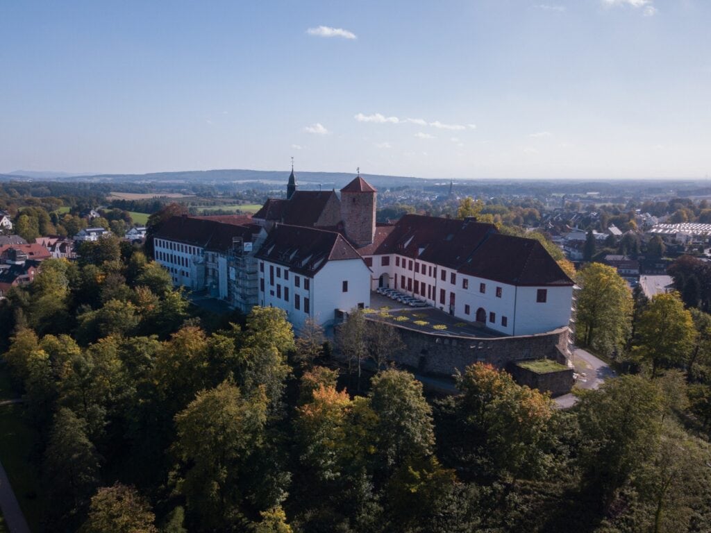 Auf einem Berg ragt eine gut sanierte Burg mit weißen, mehrstöckigen Gebäuden entlang der historischen Mauer aus einem Wald hervor. Das Land rundherum ist größtenteils bewaldet und flach, nur unmittelbar um den Burgberg herum sind die Dächer einer Stadt sichtbar. Über allem blauer Himmel.