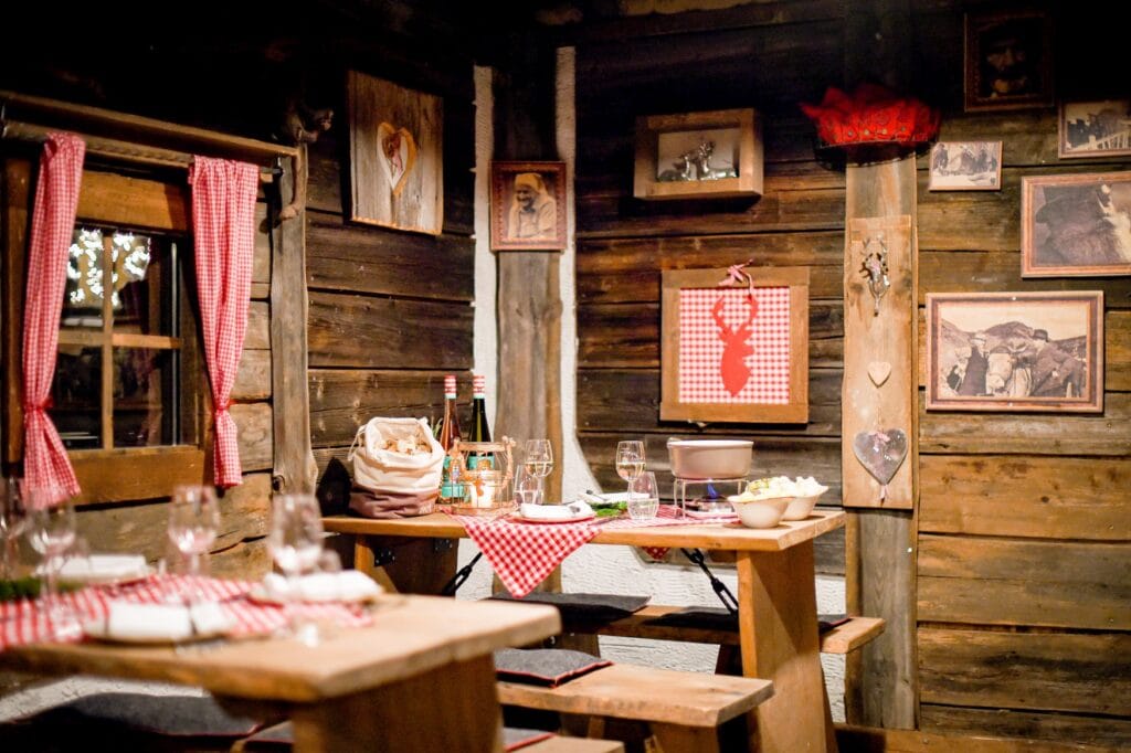 Innenraum einer typischen Almhütte als Holz; links stehen zwei hölzerne Sitzgarnituren mit rot-weiß karierten Tischdecken, auf dem hinteren Tisch dazu noch zwei Flaschen Wein und ein Fonduetopf, unter dem ein kleiner Kocher brennt. Die Wände sind mti Brettern verkleidet, daran hängen einige Schwarzweißfotos mit Alpenszenen.
