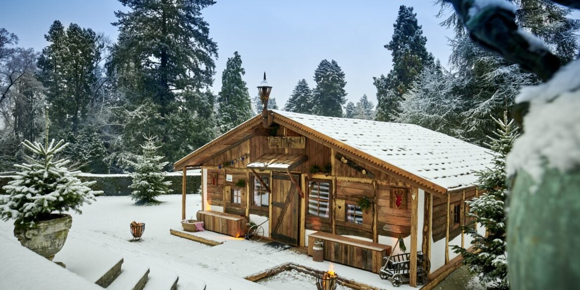 Auf einer verschneiten Gartenterrasse steht eine temporär aufgebaute Almhütte aus Holz. Alles ist verschneit. Rund um die Terrasse, zu der eine Treppe hinabführt, zieht sich eine gepflegte Hecke, dahinter rundum mehrere Bäume.